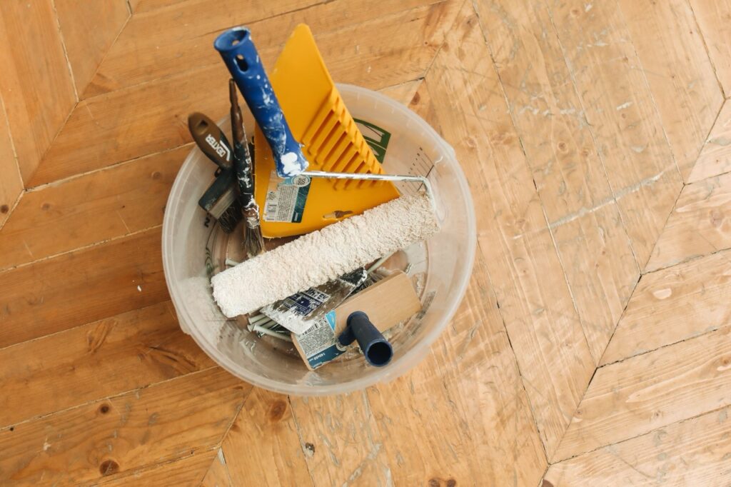 Emmer met verfroller, kwasten en plamuurmessen op een houten vloer tijdens een renovatie